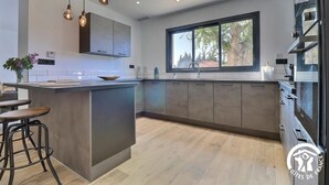 A modern kitchen with a central island, bar stools, pendant lighting, and a large window providing natural light. - Gîte Spacieux, Rénové, Climatisé Avec Grande Piscine Sécurisée, Jardin Clos (Châteaurenard)