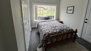 A bedroom with a bed, a window with a view of trees, a bedside table, and a framed picture on the wall. - Garden House, Unit B (Hot Springs)