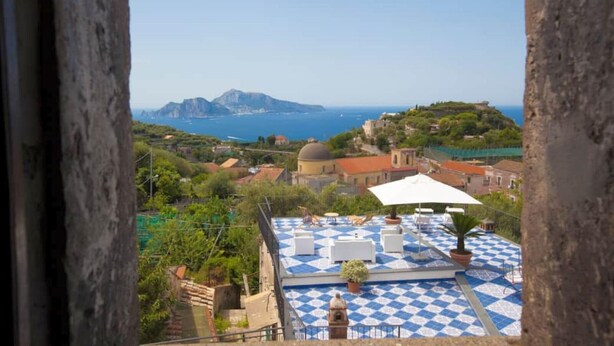 A terrace with a checkered floor, outdoor seating, and a view of the sea and distant mountains.