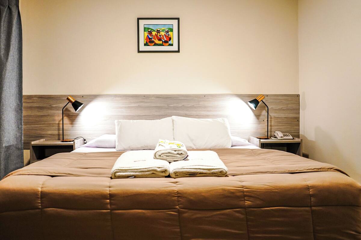 A neatly made bed with a brown bedspread, two bedside lamps, a framed picture on the wall, and a folded towel on the bed.
