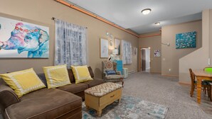 A living room with a brown sofa, yellow pillows, a wooden ottoman, and a dining table with chairs. There are artworks on the walls and a hallway leading to another room.