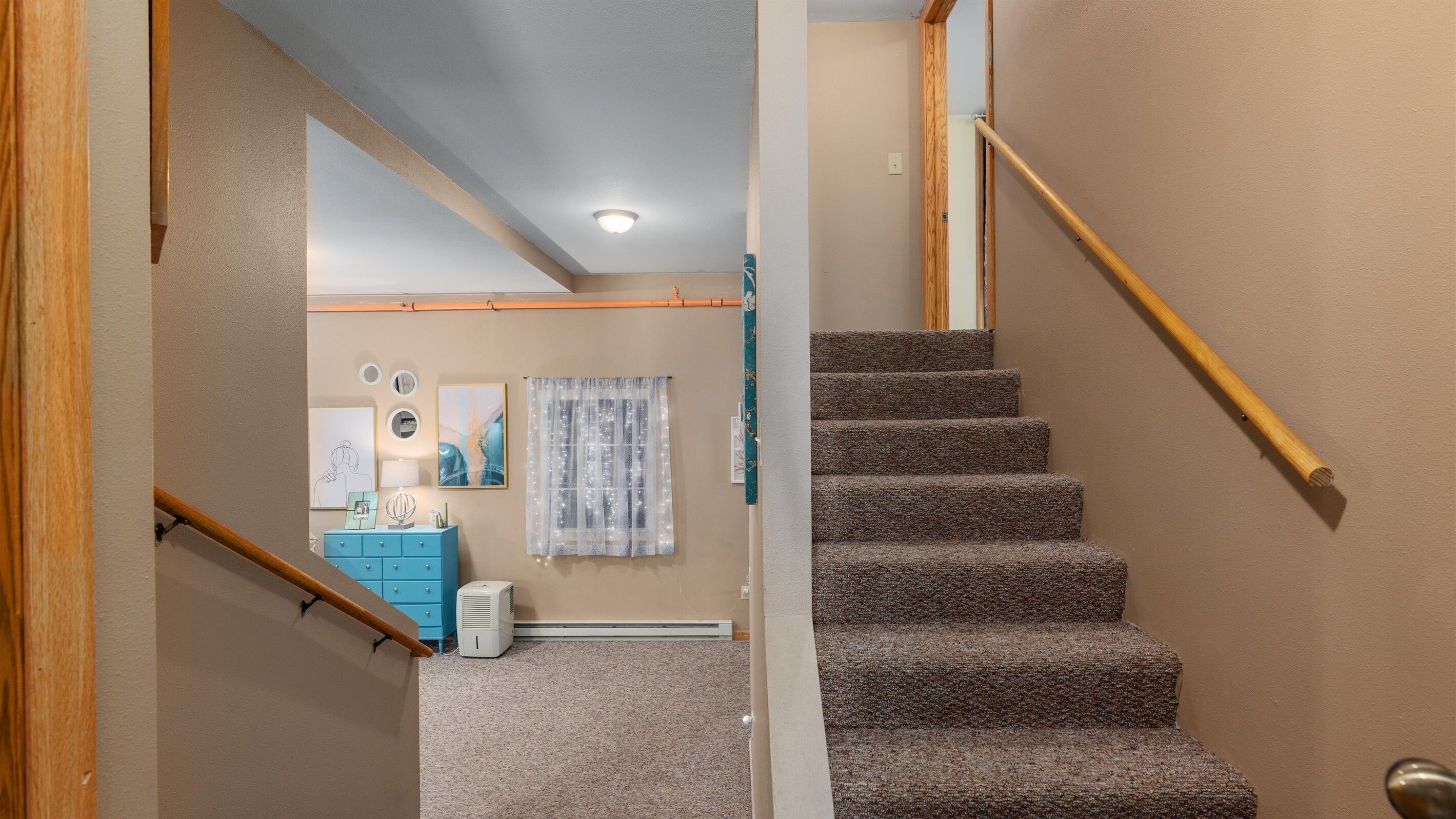 A staircase with carpeted steps and wooden handrails, leading to a room with a blue dresser, a white radiator, and wall-mounted artwork.