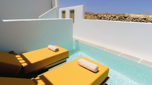 A poolside area with two yellow sun loungers, a clear blue pool, and a white wall with a stone wall in the background. -  Mr and Mrs White Paros Suites and more (Paros)