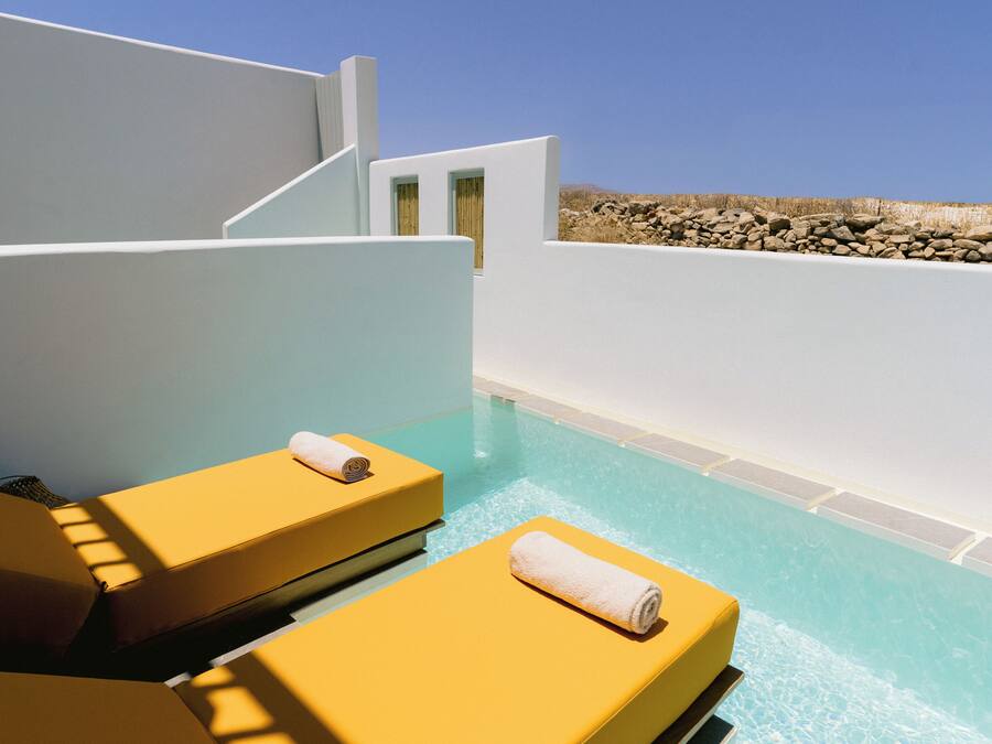 Zona junto a la piscina con dos tumbonas amarillas, una piscina azul cristalina y una pared blanca con un muro de piedra al fondo.