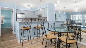 Une cuisine moderne avec une table à manger en verre et des tabourets de bar, des murs bleu clair et un réfrigérateur bien visible.