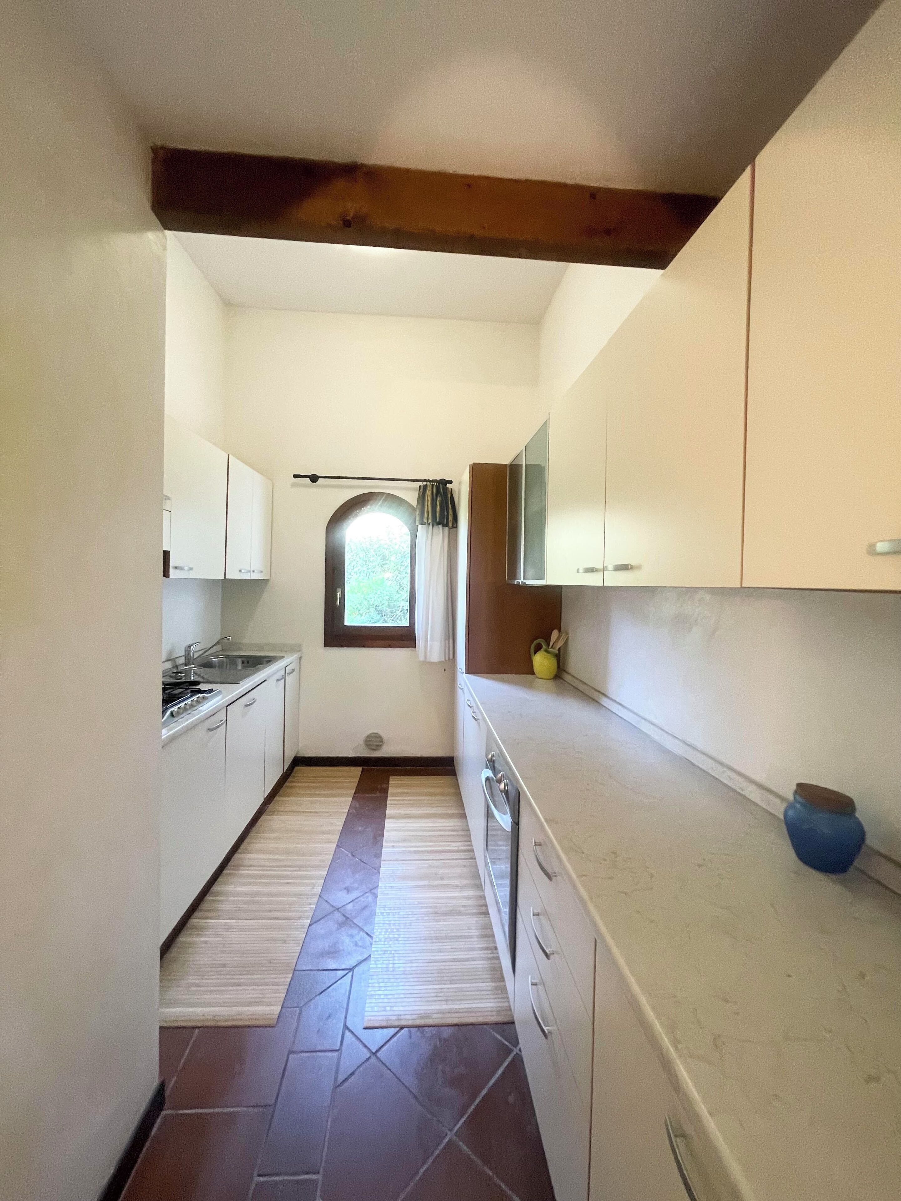 A narrow kitchen with white cabinets, a tiled floor, and a window with curtains.