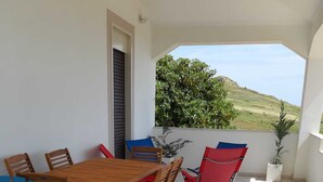 A covered outdoor dining area with a wooden table and chairs, a potted plant, and a view of a hill.