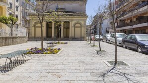 A paved walkway with benches and flower beds, flanked by parked cars and buildings.