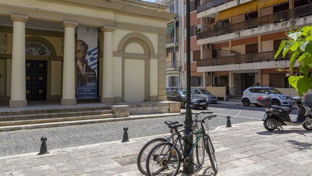 A building with classical architecture, including columns and a large arched doorway. There is a banner with text and a flag on the building's facade.