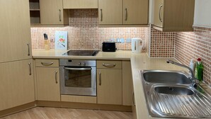 Een kleine keuken met houten kastjes, een inbouwoven, een magnetron, een waterkoker en een spoelbak met een roestvrijstalen kom.