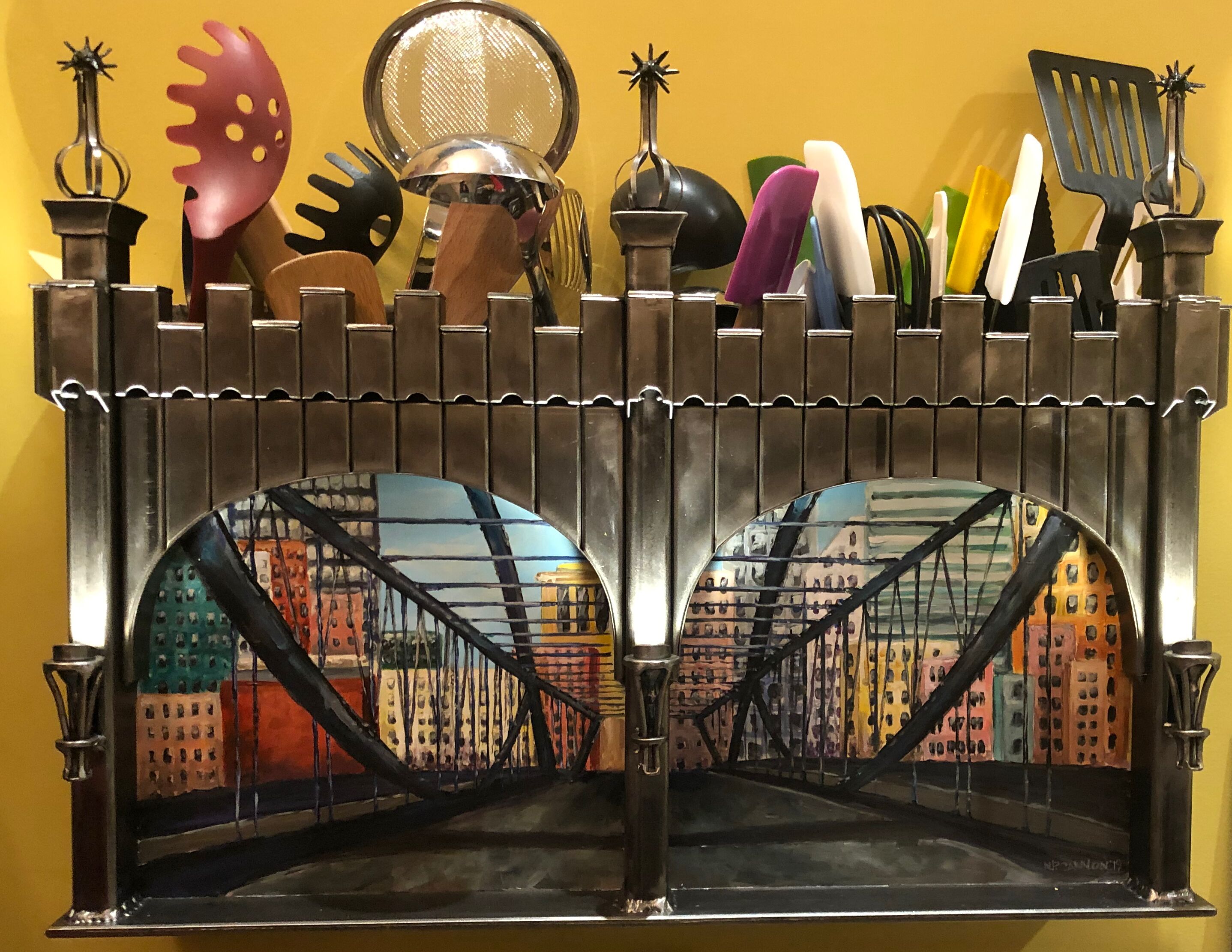 A metal fence with a painted cityscape mural, a mirror, and various kitchen utensils.