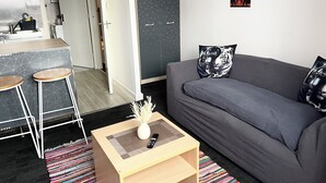 En moderne stue med en grå sofa, to matchende pude med tiger-motiv, et træbord med vase og fjernbetjening, og et køkkenområde med en barstol og flisebeklædt væg.