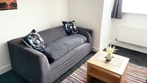 En moderne stue med en grå sofa, to puder med mønster, et lille træsidebord med en vase og en farverig stribet måtte.
