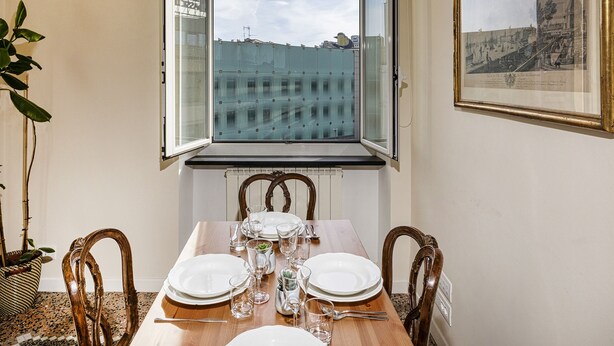 Uma área de refeições com uma mesa posta para quatro pessoas, uma janela com vista para edifícios e um quadro na parede.