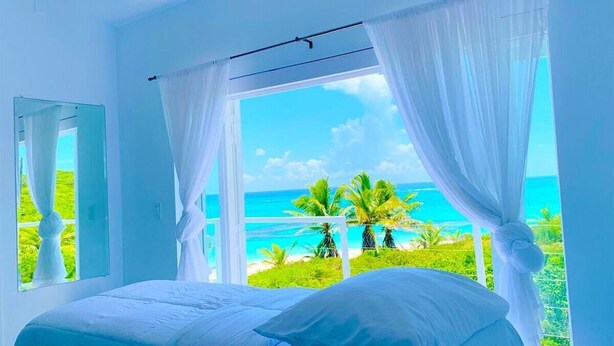 Een slaapkamer met een bed, witte gordijnen en uitzicht op een strand en palmbomen.