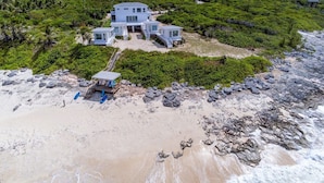 Une propriété située en bord de mer, dotée d’une maison moderne, d’un espace couvert avec des sièges et d’un escalier menant à la plage.