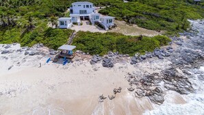 Una propiedad frente al mar con una casa moderna, un espacio cubierto con asientos y una escalera que lleva a la playa.