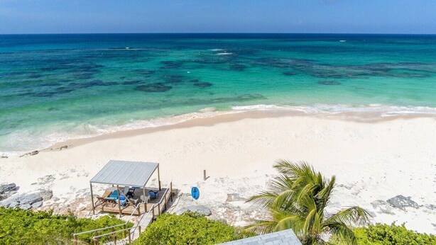 Una playa con agua turquesa cristalina, arena blanca y una pequeña estructura con sombra y asientos.