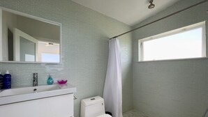 Un baño con inodoro blanco, ducha con cortina blanca, un espejo grande sobre el lavamanos y una ventana que proporciona luz natural.