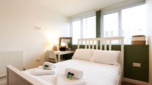 Ein ordentlich bezogenes Bett mit weißen Leinen und Handtüchern, ein Nachttisch mit Lampe, ein Sessel und ein Fenster mit Jalousien.
