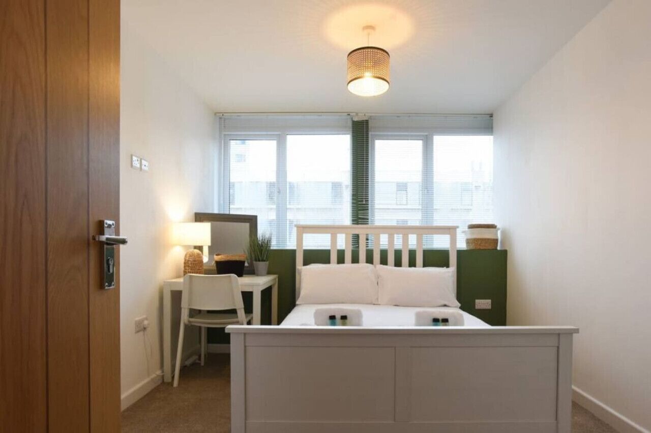 A bedroom with a bed, a desk, a chair, a lamp, and a window.