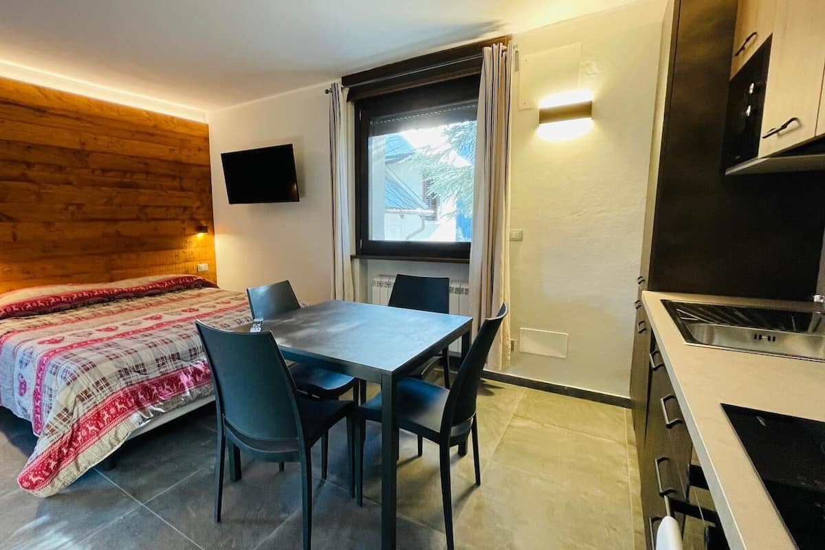 A compact room with a bed, a dining table with chairs, a TV mounted on the wall, and a kitchenette.
