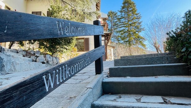 A wooden welcome sign with the word "Welcome" and "Willkommen" in English and German, respectively, above a set of stone steps leading to a building. - Residence VILLA LINDA (Bardonecchia)