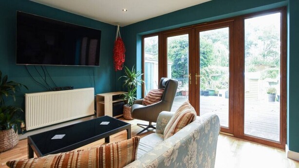 A living room with a sofa, armchair, and coffee table. There is a TV mounted on the wall and a window with a view of greenery outside. - The Nest | Large Family Home! (Cowplain)
