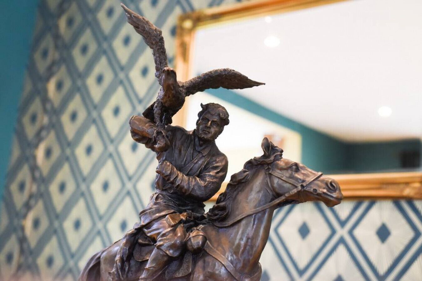 Een bronzen beeld van een ruiter met een adelaar, voor een muur met een patroon.