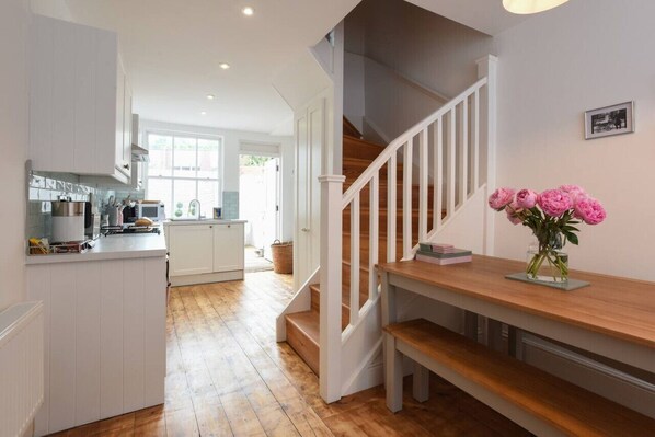 A modern kitchen with white cabinets and a wooden floor. A staircase with white railings leads to a landing with a vase of pink flowers.