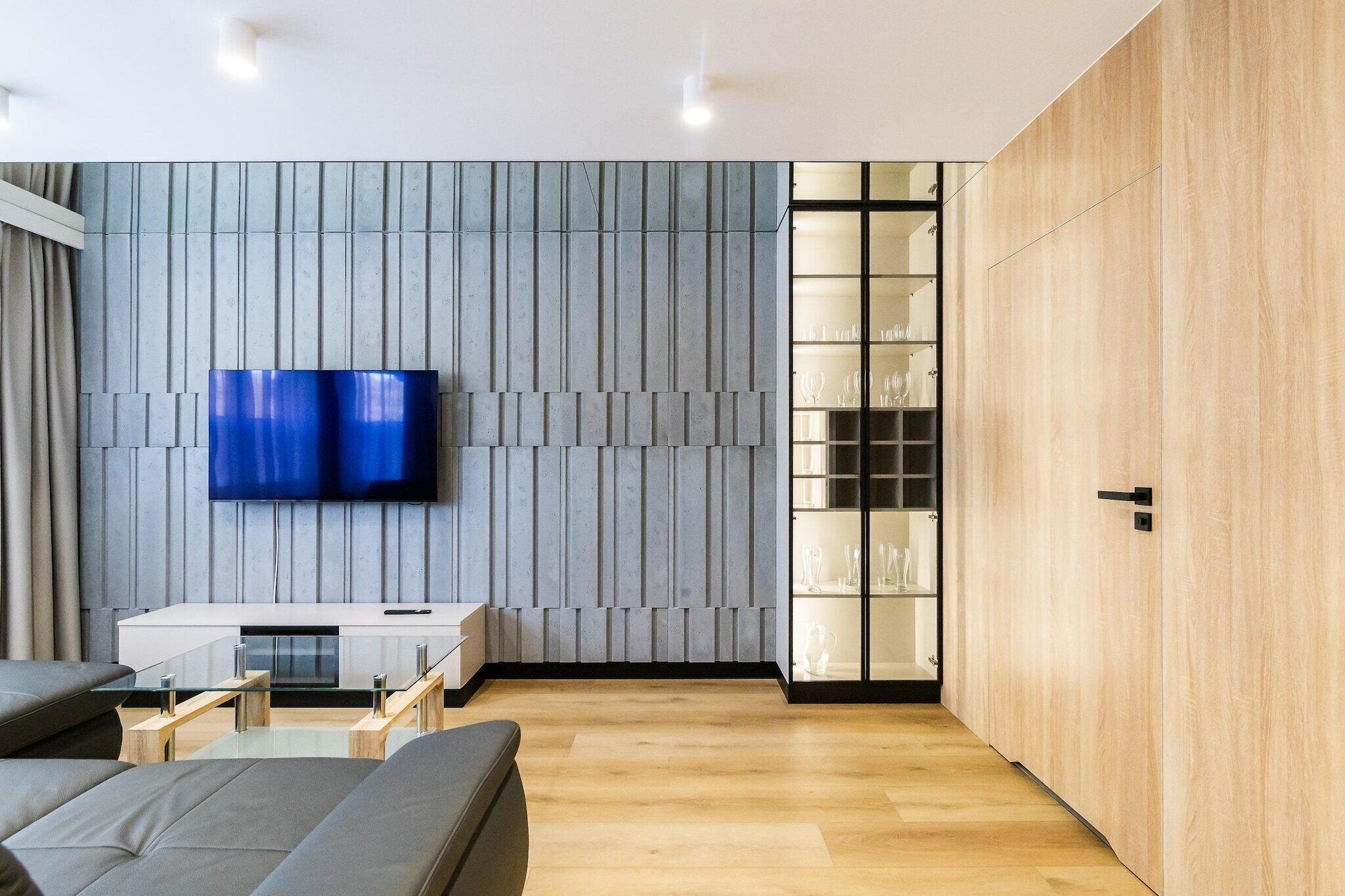 A modern living room with a flat-screen TV mounted on a textured wall, a wooden cabinet, and a glass coffee table.