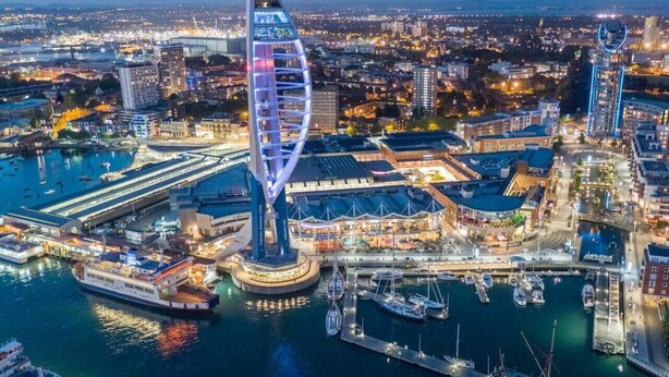 A modern, illuminated building with a unique spiral design, situated in a bustling urban waterfront area with a marina and various buildings around.