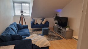 Una sala de estar moderna con un sofá azul, un sillón azul más pequeño, un televisor sobre un mueble gris y un trípode en la esquina.