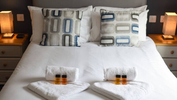 A neatly made bed with white linens, two pillows with geometric patterns, and two towels with orange and black containers on them. - Parkside Haven | 2BR w/Off-Road Parking (Hampshire)