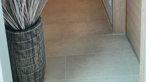 Un pasillo con una puerta de madera a la derecha, un accesorio de luz en el techo y una planta con flores blancas y púrpuras a la izquierda.