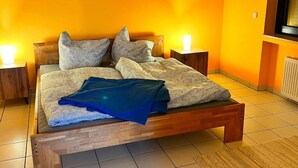 A bed with a wooden frame, two pillows, and a blanket. There are two bedside tables with lamps. The room has orange walls and a tiled floor. - Zeitwohnung Kenn/region Trier (Kenn)