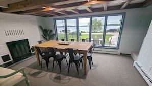 A dining area with a wooden table and chairs, a fireplace, and a large window with a view of a lake and vehicles. - Charming 4-bedroom Lake house on Lake Ripley in Cambridge (Cambridge)