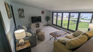 A living room with a sofa, a coffee table, a TV on a cabinet, and large windows with a view of a body of water and a dock. - Charming 4-bedroom Lake house on Lake Ripley in Cambridge (Cambridge)