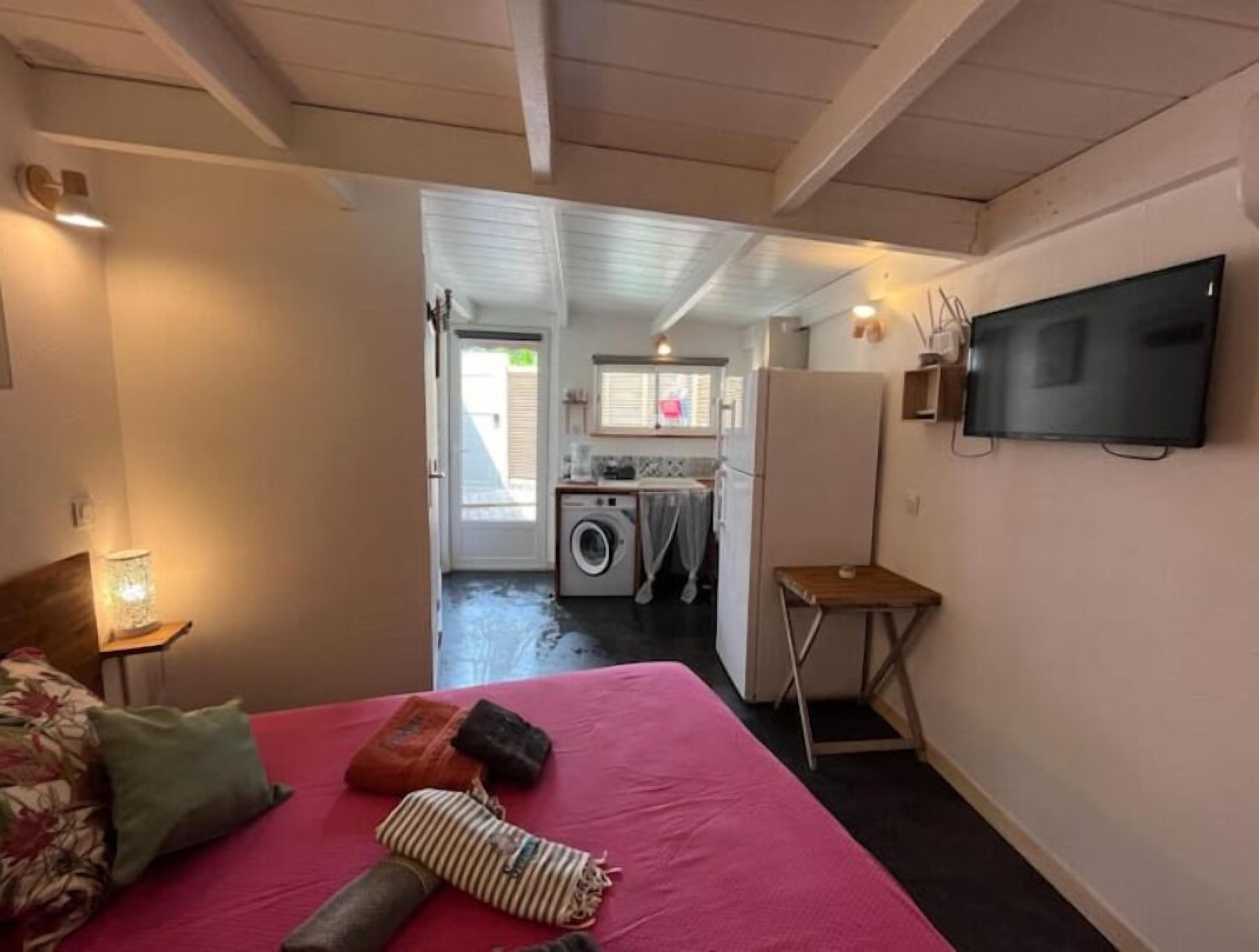 A bedroom with a bed, a small table, a TV, and a view into the kitchen area.