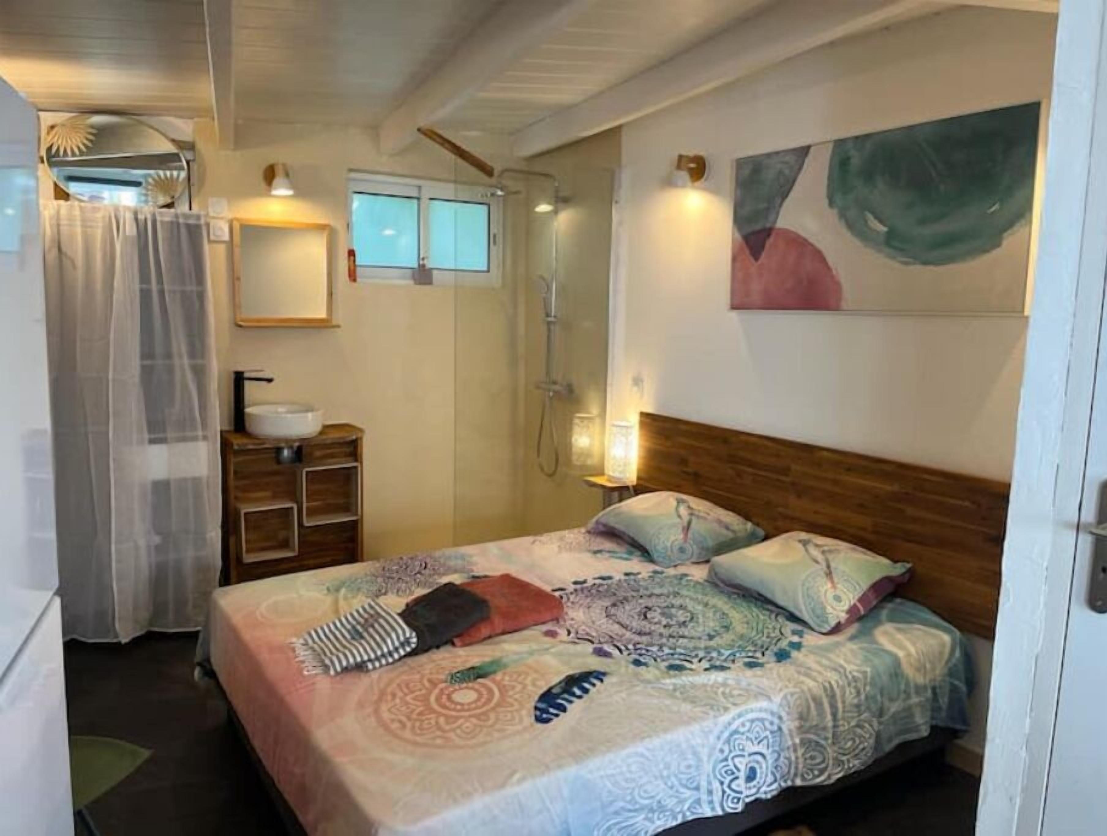 A bedroom with a bed, a bedside table, a mirror, a shower, and a window.