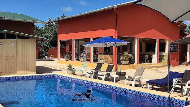 A swimming pool with lounge chairs and umbrellas, a red building with an open patio, and a sign reading "RESIDENCE AINA". - RESIDENCE AINA (Abomey-Calavi)