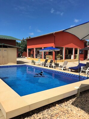 Una piscina con sillas de descanso y sombrillas, un edificio rojo con un patio abierto y un letrero que dice "RESIDENCE AINA".