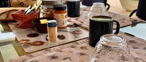 Una mesa dispuesta con diversos alimentos, una taza de café y un tarro de vidrio, probablemente en un comedor con vistas al exterior.