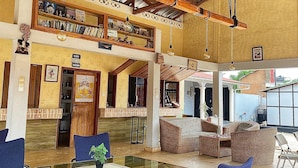 A spacious indoor area with a high ceiling and wooden beams, featuring a central glass table surrounded by blue chairs, and a lower level with wicker furniture and a bar area. - RESIDENCE AINA (Abomey-Calavi)