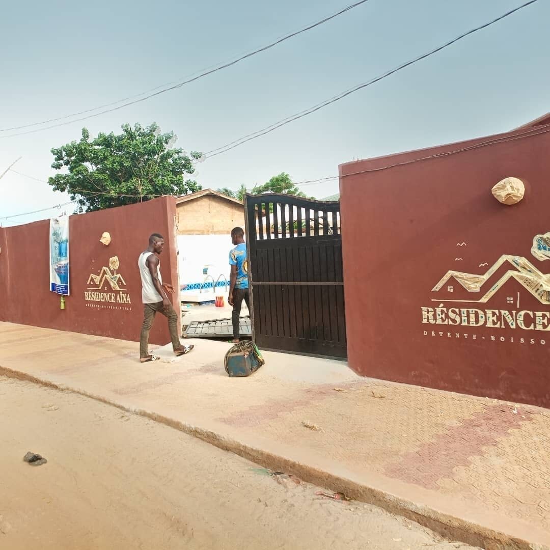 Una entrada protegida por un muro color borgoña y un portón negro, con el logo 'Résidence Aïna' y la frase 'Détenue - Boisso'.