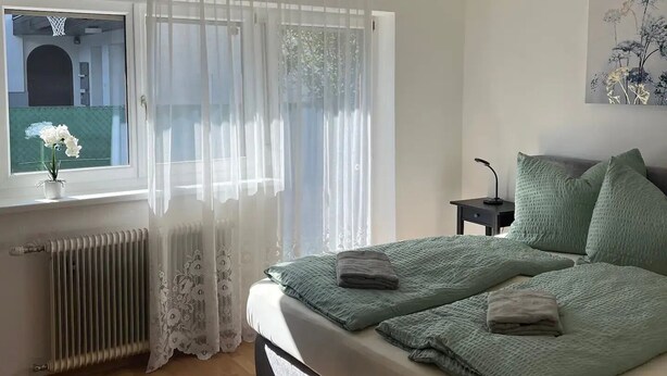 A bedroom with a large bed, a radiator, a window with sheer curtains, a small table with a lamp, and a painting on the wall. - Apartment mit Gartenblick (Aldrans)