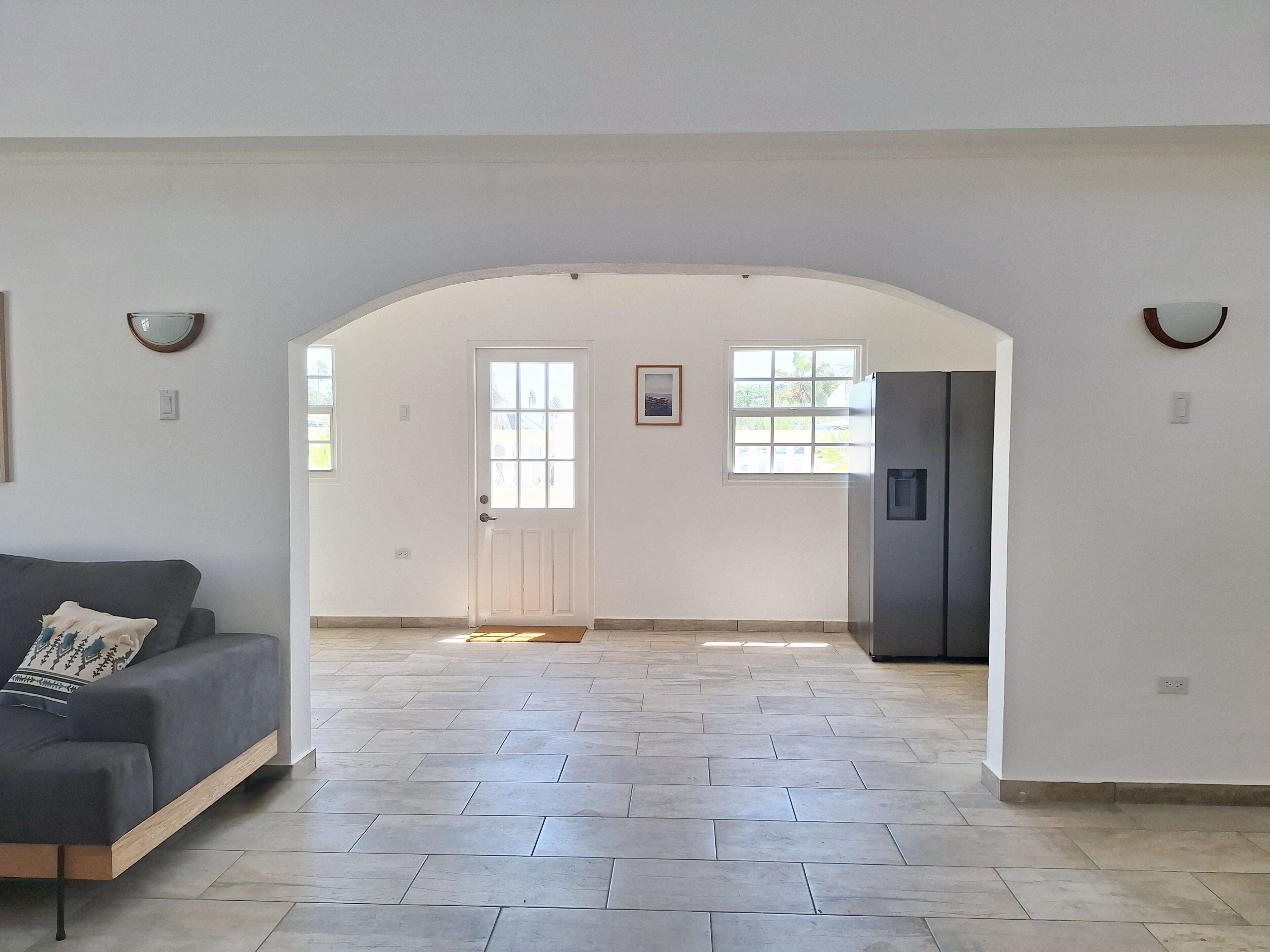 Amplia sala de estar con un sofá gris, una puerta que da a otra habitación y un ventanal grande.