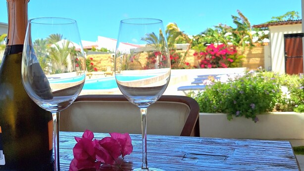 A wooden table with two wine glasses and a bottle of wine, set against a view of a swimming pool and greenery. - NEW! Villa La Brisa Aruba│Beautiful&Relaxing: Private pool,8-Min Stroll to Beach (Noord)