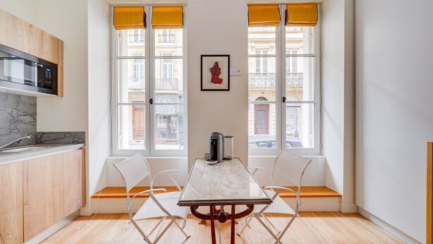 Una cocina moderna con microondas integrado, armarios de madera y encimera de mármol. Hay dos ventanas con persianas amarillas, un cuadro enmarcado en la pared y una mesa con sillas en el centro.