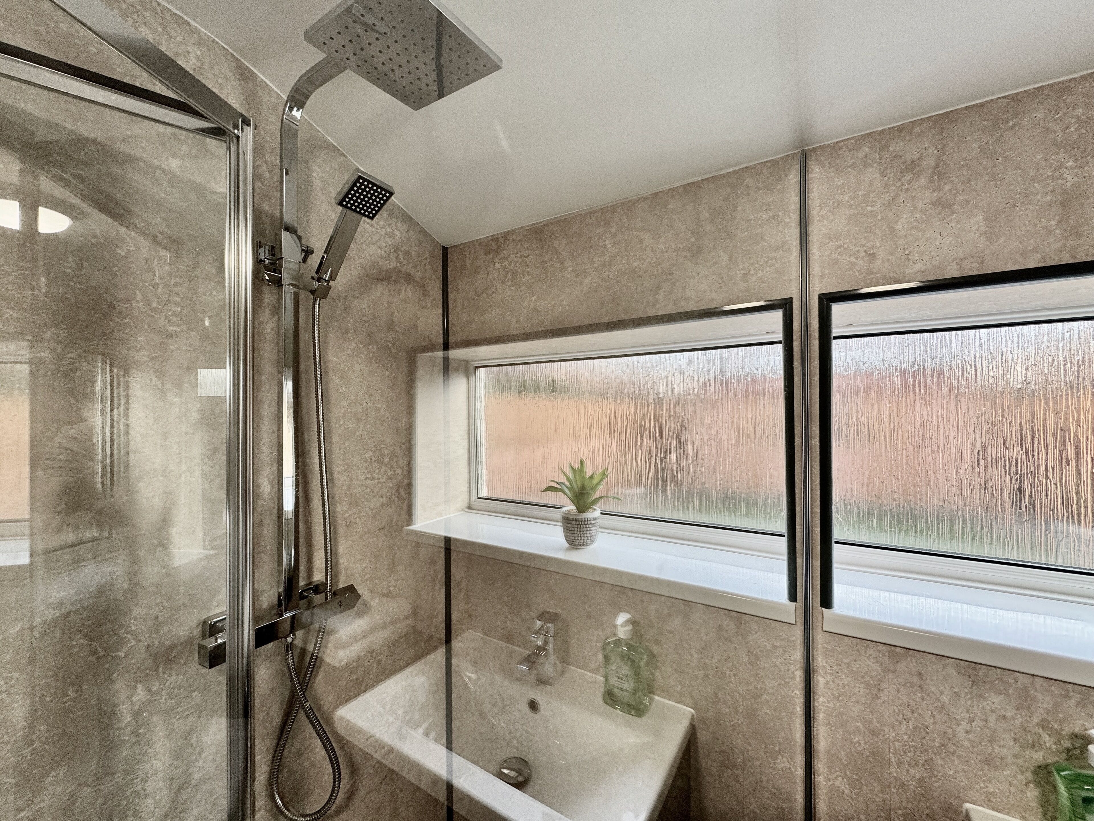 A modern bathroom with a glass-enclosed shower, a sink with a window above it, and a small shelf with a plant and toiletries.